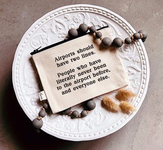 A lifestyle picture of the natural canvas zipper pouch with a black YKK zipper, printed in black with "Airports should have two lines. People who have literally never been to the airport before, and everyone else."