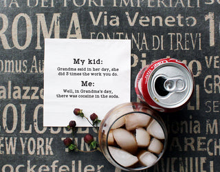 A lifestyle shot of a stack of a cocktail napkin printed in black with "Grandma said in her day, she did 3 times the work you do. Me: Well, in grandma's day, there was cocaine in the soda." Shown with a can of coke and a glass of coca-cola on ice.
