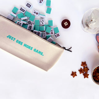 Canvas pouch with 'Just One More Game' text, mahjong pieces, and snacks on a white surface
