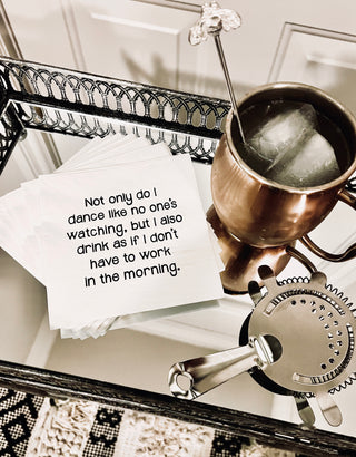 A lifestyle picture of a stack of white 3-ply cocktail napkin printed in black with "Not only do I dance like nobody is watching but I also drink as if I don't have to work in the morning." shown with a cocktail and a strainer on a table.
