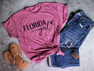 Raspberry t-shirt with 'Florida Girl' text, denim shorts, and sandals on a gray surface