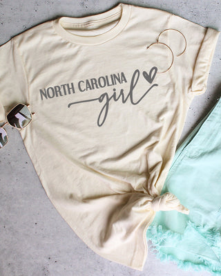 Beige t-shirt with 'North Carolina girl' text, sunglasses, and light blue shorts on a gray surface