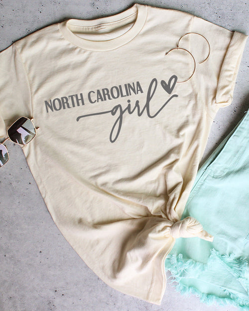 Beige t-shirt with 'North Carolina girl' text, sunglasses, and light blue shorts on a gray surface