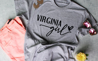 Light gray tee shirt with 'Virginia girl' text on a gray surface with pink pants and sunglasses.
