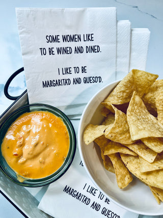 Lifestyle shot of white 3-ply cocktail napkins printed in black with "Some women like to be wined and dined. I like to be margarita'd and queso'd." Shown with a bowl of tortilla chips and cheese.