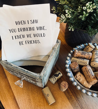 A lifestyle shot of a stack of white 3-ply cocktail napkins printed in black with "When I saw you drinking wine I knew we would be friends." Shown with a bowl of wine corks and a plant on a table. 