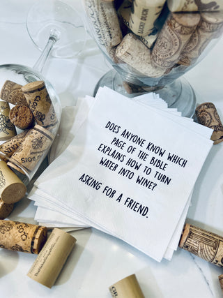 A lifestyle shot of a stack of white 3-ply cocktail napkins printed in black with "Does anyone know which page of the Bible explains how to turn water into wine? Asking for a friend." Shown with a bunch of wine corks.