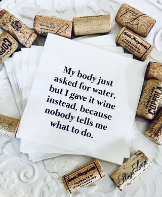 A lifestyle shot of a stack of white 3-ply cocktail napkins printed in black with "My body asked for water, but I gave it wine instead, because nobody tells me what to do." Shown with wine corks on a fancy white plate.