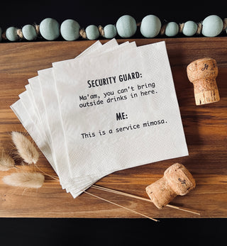 A lifestyle shot of a stack of white 3-ply cocktail napkins printed in black with "Security Guard: Ma'am, you can't bring outside drinks in here. Me: This is a service mimosa." shown with 2 champagne corks and  a string of beads.